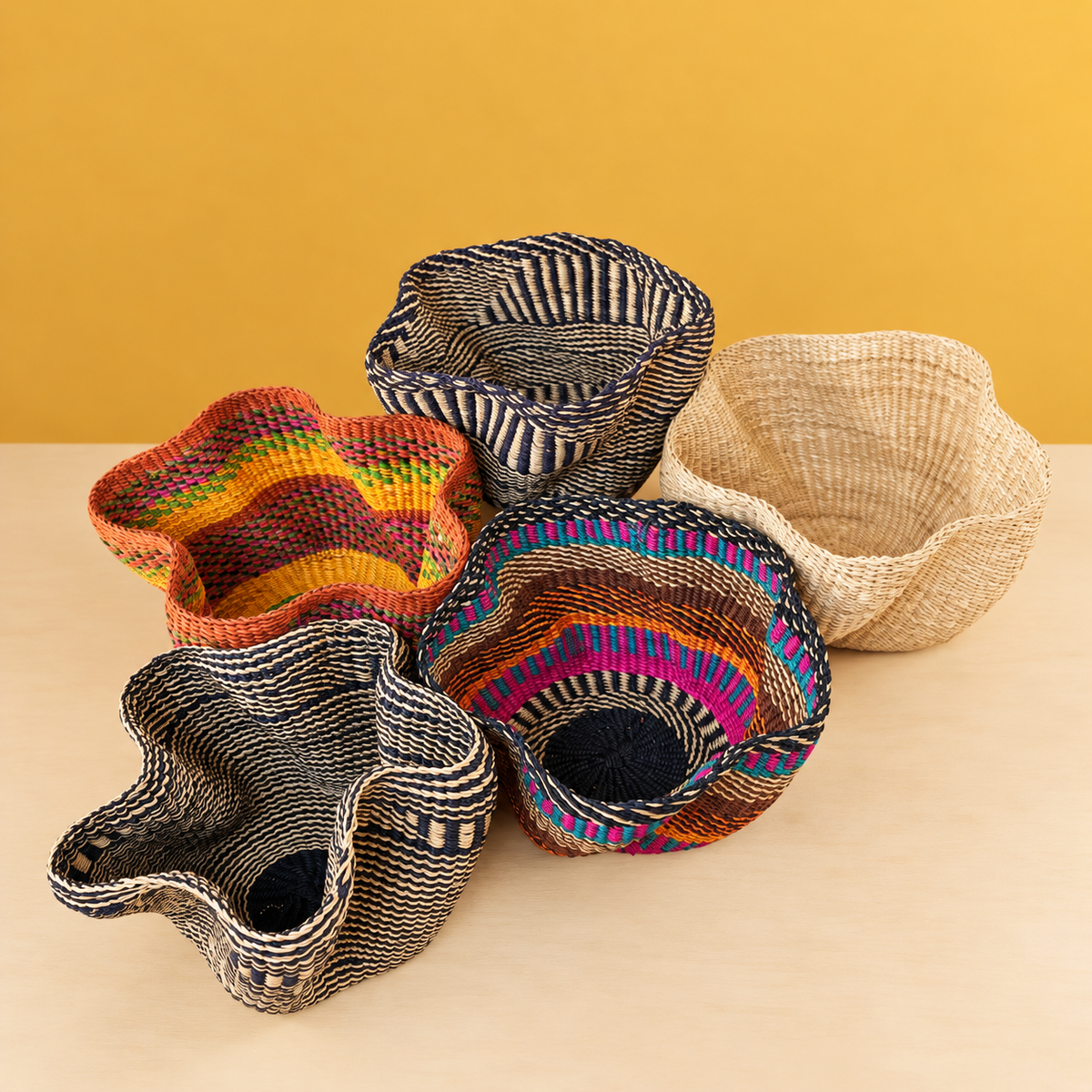 Five handwoven baskets with wavy rims in bold colours and natural straw, on a mustard backdrop
